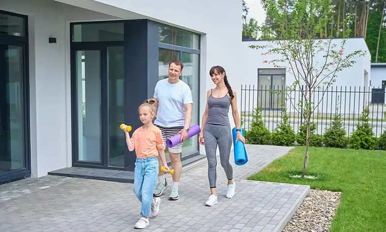 A family walks outside a modern house, with the child carrying dumbbells and the adults holding yoga mats.