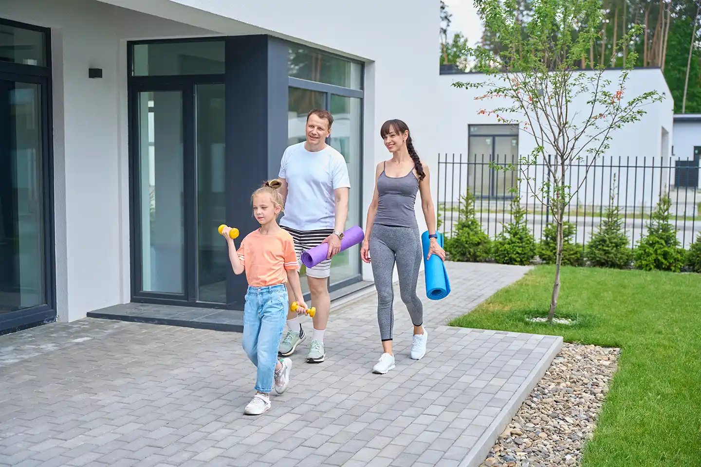 A family walks outside a modern house, with the child carrying dumbbells and the adults holding yoga mats.