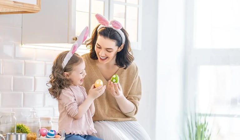Parent and child sitting together in a bright kitchen, wearing bunny ear headbands and holding decorated Easter eggs