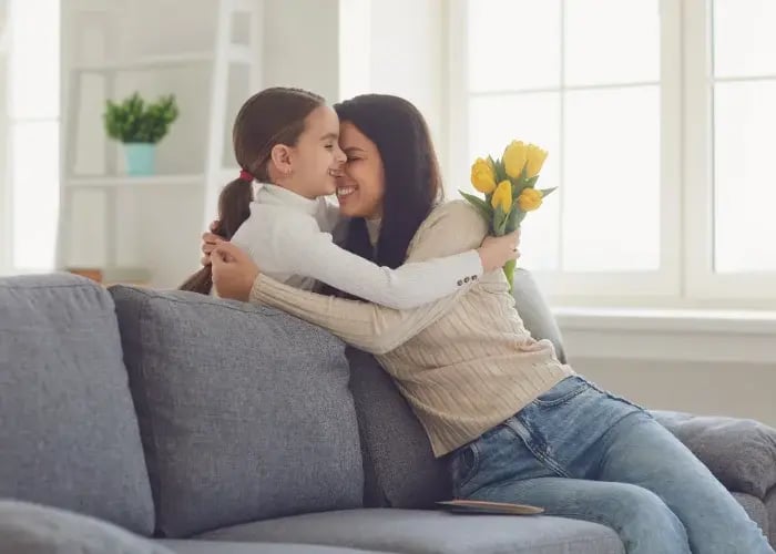 Parent and child sitting on a sofa, sharing a hug while the parent holds a small bouquet of yellow tulips in a bright living room
