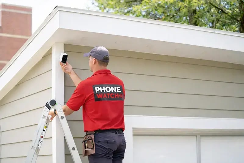 PhoneWatch security advisor installing a wireless battery video camera on a garage wall using a ladder and tools
