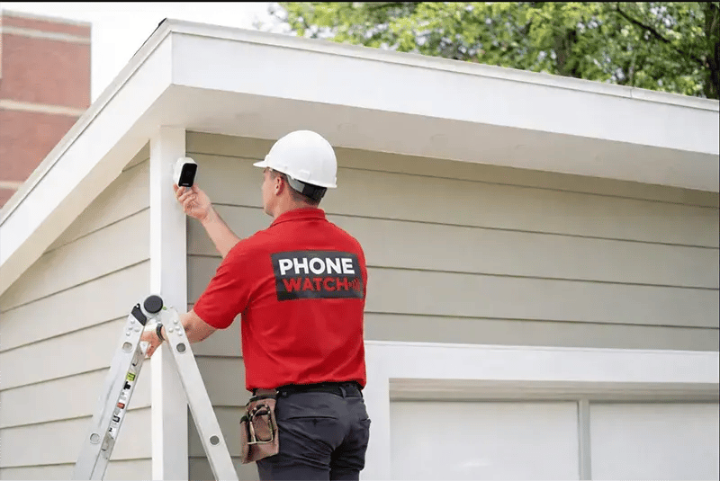 PhoneWatch security advisor installing a wireless battery video camera on a garage wall using a ladder and tools