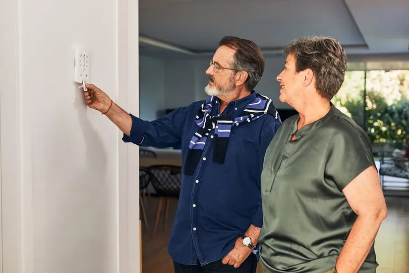 two people entering a code on a wall mounted home alarm keypad while another person stands nearby inside a bright modern home.